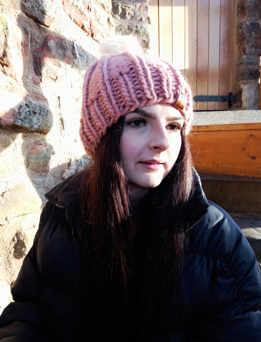 This image shows a young woman with long dark hair and black coat wearing a chunky hand knitted hat in pink, featuring a creamy coloured pompom. 