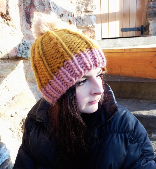 This image shows a super chunky knitted hat in mustard with a pink brim, worn by a young lady with long dark hair. It features a fluffy creamy coloured pompom. 