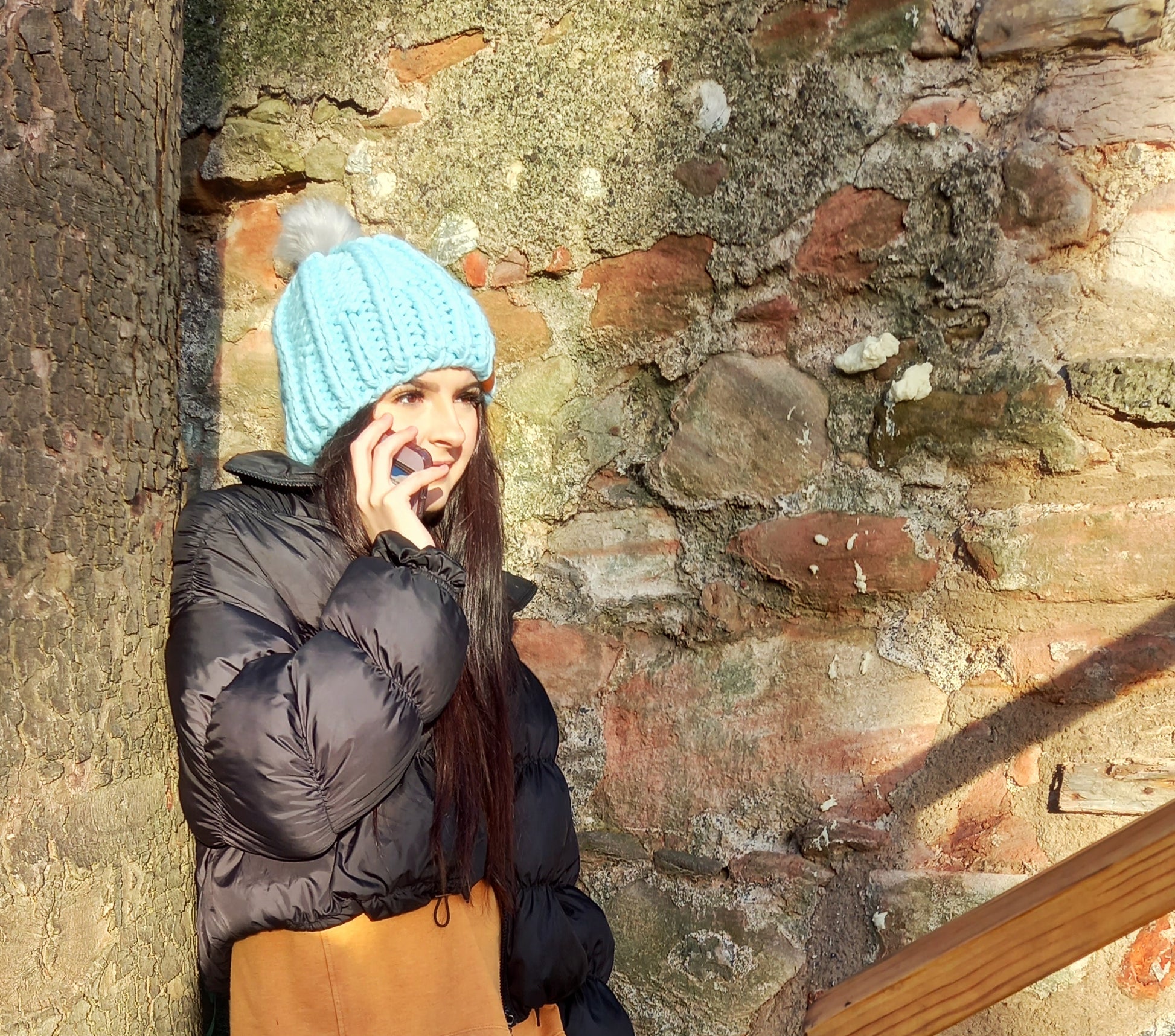 This image shows a young lady with long dark hair talking on a mobile phone. She is wearing a baby blue super chunky handknitted hat featuring a light grey pompom.