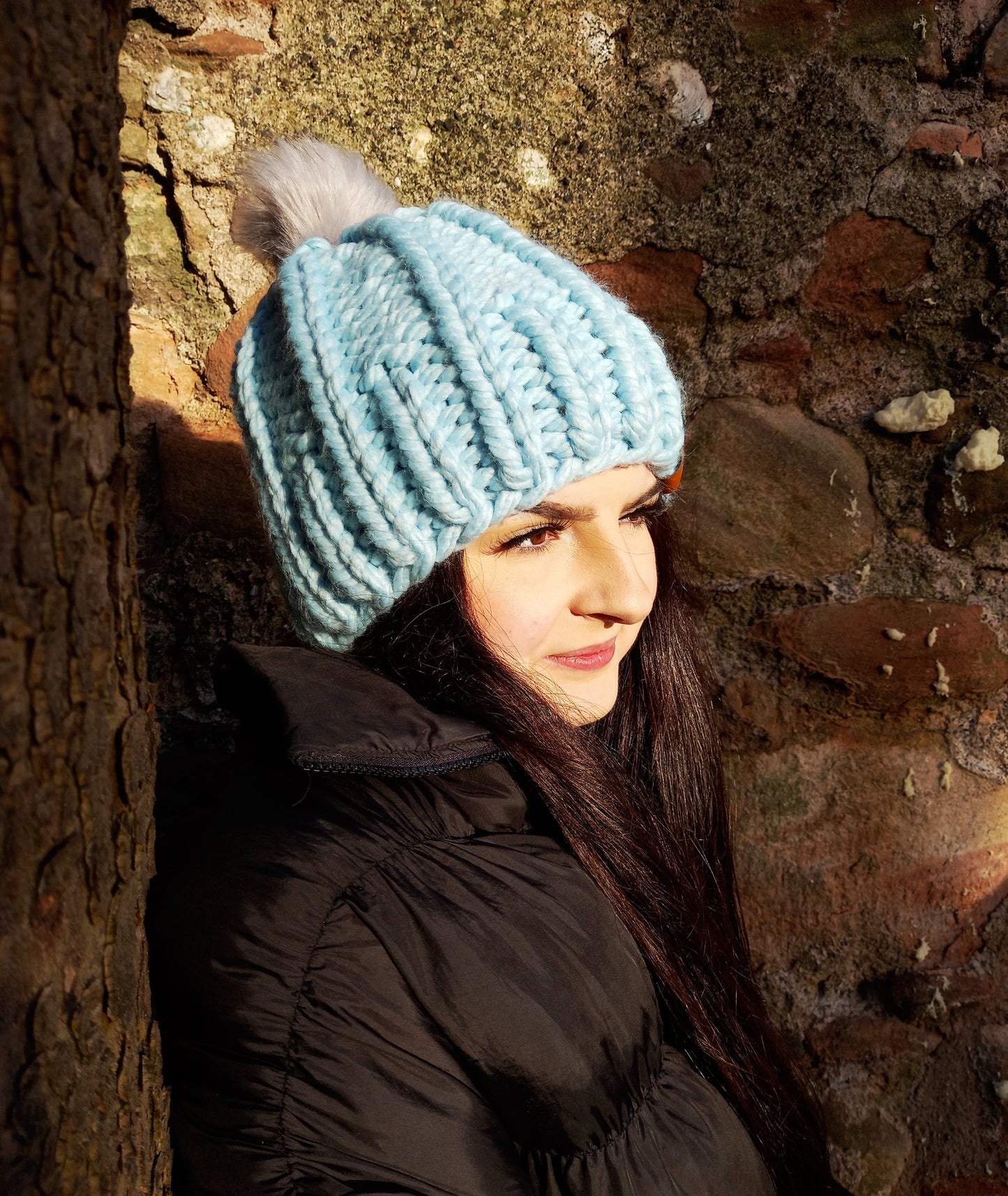 This image shows a young lady with long dark hair wearing a baby blue super chunky handknitted hat featuring a light grey pompom.