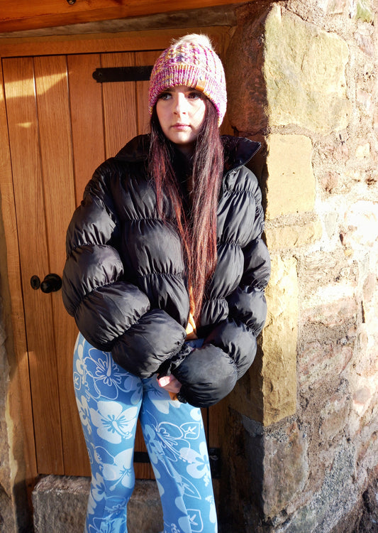 This image shows a young lady with long dark hair, standing in a doorway, wearing a hand knitted hat with a faux cable pattern with a wide fold up brim and is a lovely mixture of pinks, yellows, purples and blues. It features a small faux leather 'Taffeta Fox' label and a soft cream removeable pompom.