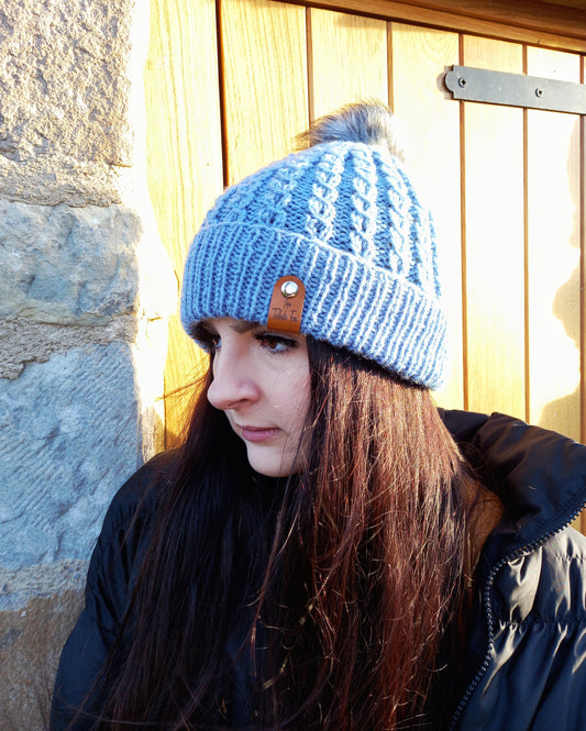 This image shows a young lady with long dark hair wearing a faux cable hand knitted hat with turned up brim in a lovely shade of blue. The hat features a light grey pompom and brown faux leather The Taffeta Fox tag.