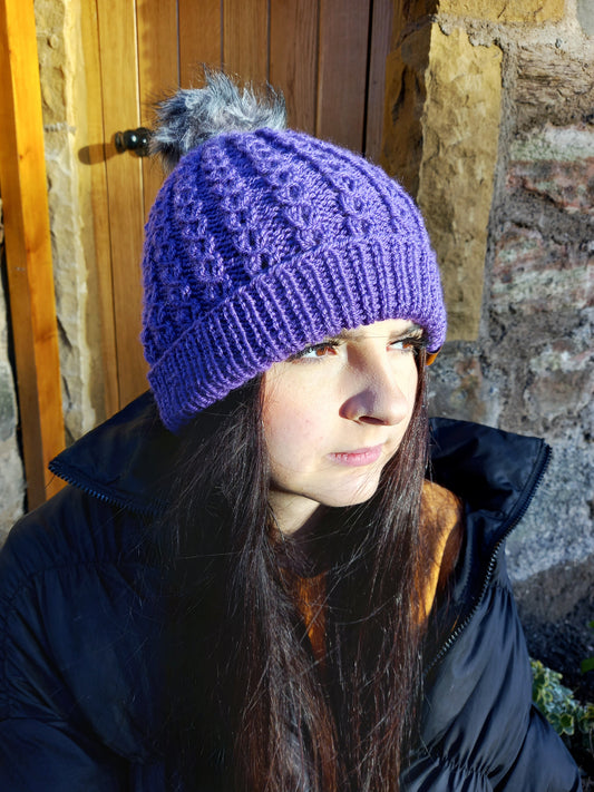 This image shows a young woman with long dark hair wearing a bright purple hand knitted faux cable hat featuring a light grey pompom.