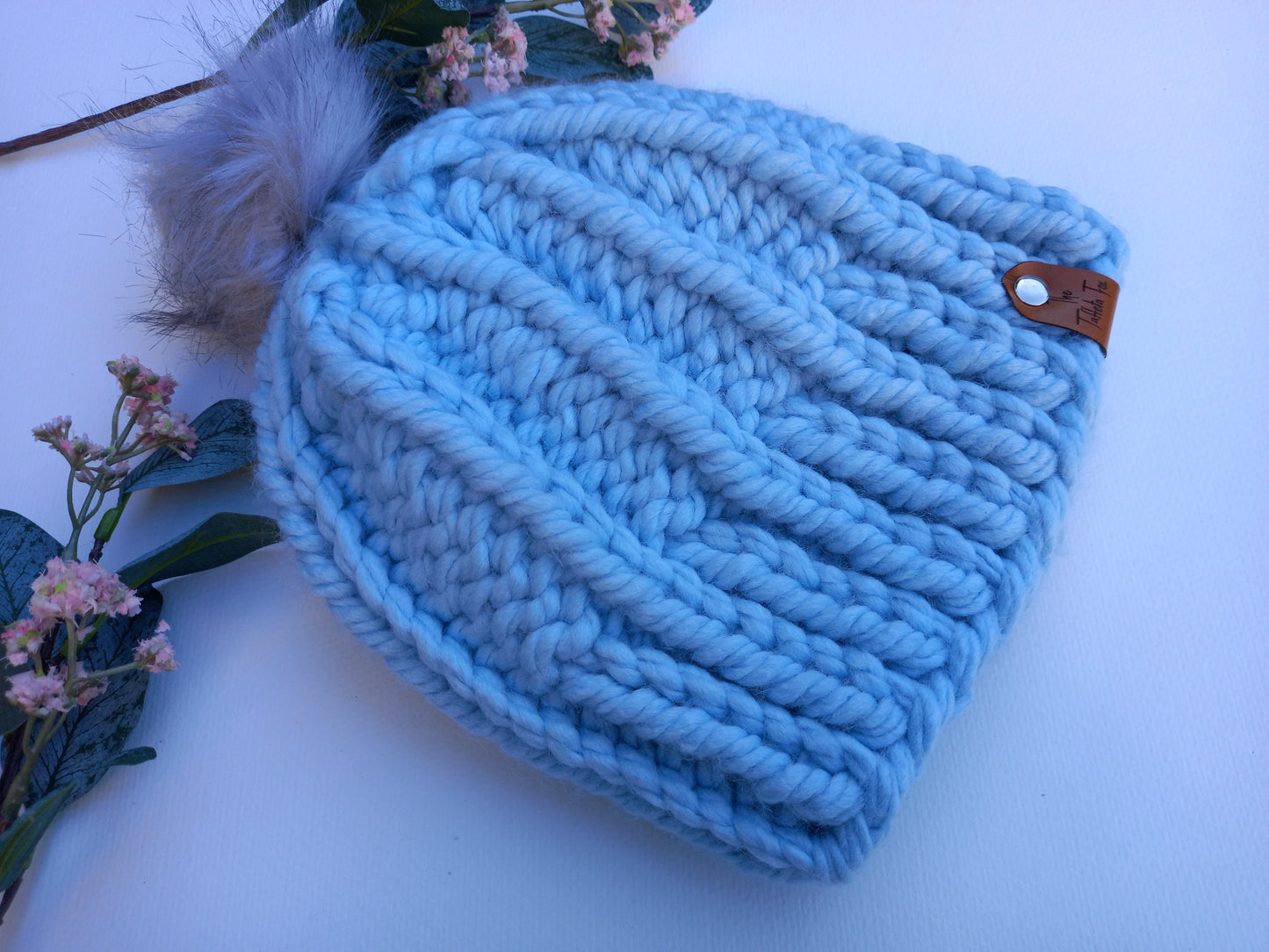 This shows a baby blue super chunky hand knitted hat lying on a white background with some foliage and pink floral decoration. 